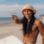 A smiling person with long hair and a hat holds up a tin of Badger Balm Adventure Mineral Sunscreen Tin - SPF 50 on a sandy beach, with the ocean and a surfboard in the background. Its zinc oxide formula makes it reef friendly, perfect for sunny days by the water.
