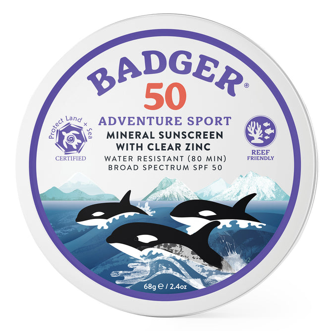 Badger Balm Adventure Mineral Sunscreen Tin - SPF 50 comes in a round tin featuring clear zinc oxide. The Adventure Sport label highlights three orcas swimming amid mountains and ocean illustrated in purple, blue, and white. Reef friendly and adorned with certified symbols, this sunscreen provides mineral protection.