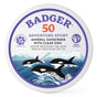 Badger Balm Adventure Mineral Sunscreen Tin - SPF 50 comes in a round tin featuring clear zinc oxide. The Adventure Sport label highlights three orcas swimming amid mountains and ocean illustrated in purple, blue, and white. Reef friendly and adorned with certified symbols, this sunscreen provides mineral protection.