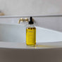 A glass dropper bottle of yellow oil labeled TERRA sits on the edge of a white bathtub, with a blurred bathtub faucet in the background.