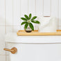 A white toilet tank with a wooden tray supports a small potted plant and a roll of Grove Co. 3-Ply Bamboo Toilet Paper. The wall is adorned with white tiles, and the flush handle is metallic.