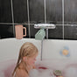 A young blonde-haired child sits in a pink, bubbly bath. On the tub’s edge is a mushie Bath Rinse Cup – Soft Silicone, Easy-Grip; nearby are a blue faucet cover and two floating cloud-shaped toys against a black-tiled wall.