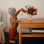 A toddler in a gray hat and brown jacket stands by a wooden table, reaching for a pink mushie Bath Rinse Cup – Soft Silicone, Easy-Grip, filled with colorful flowers. Books are under the table, and a light-colored sofa sits nearby.