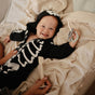 A smiling baby in a black skeleton onesie lies on a bed, holding a toy. In the foreground, an adult uses the mushie Diaper Cream Applicator – Soft Silicone, Mess-Free—perfect for your list of baby essentials.