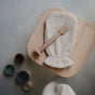 A beige exfoliating mitt and the mushie Diaper Cream Applicator – Soft Silicone, Mess-Free rest on a light wooden stool; small baby essentials are scattered out of focus on the gray floor below.