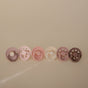 Six mushie FRIGG Natural Rubber Pacifiers for ages 0-6 months in pastel purple, pink, cream, and brown are lined up on a beige background. Each Denmark-made, BPA-free pacifier features a scalloped edge and circular handle.