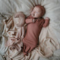 A newborn sleeps on cream bedding, wearing a rust outfit and hat, soothing with a mushie FRIGG Natural Rubber Pacifier (BPA-Free, Baby Safe, 0-6 months); a crescent moon-shaped pillow rests beside them.
