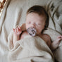 A baby sleeps peacefully with the mushie FRIGG Natural Rubber Pacifier—BPA-Free and safe for 0-6 months—under a cream, dotted blanket. The natural rubber soothes, their light brown hair and calm face completing the serene scene.