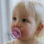 A young toddler with blonde hair and blue eyes looks upward, holding a mushie FRIGG Natural Rubber Pacifier – BPA-Free 6-Pack in light purple. Soft natural light filters in from the background.