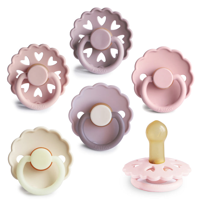 Six pastel FRIGG Natural Rubber Pacifiers by mushie, BPA-free 6-pack, are arranged in two rows. One pacifier displays its nipple side view. Colors shown include soft pink, lavender, and cream; each features scalloped edges and heart cutouts.