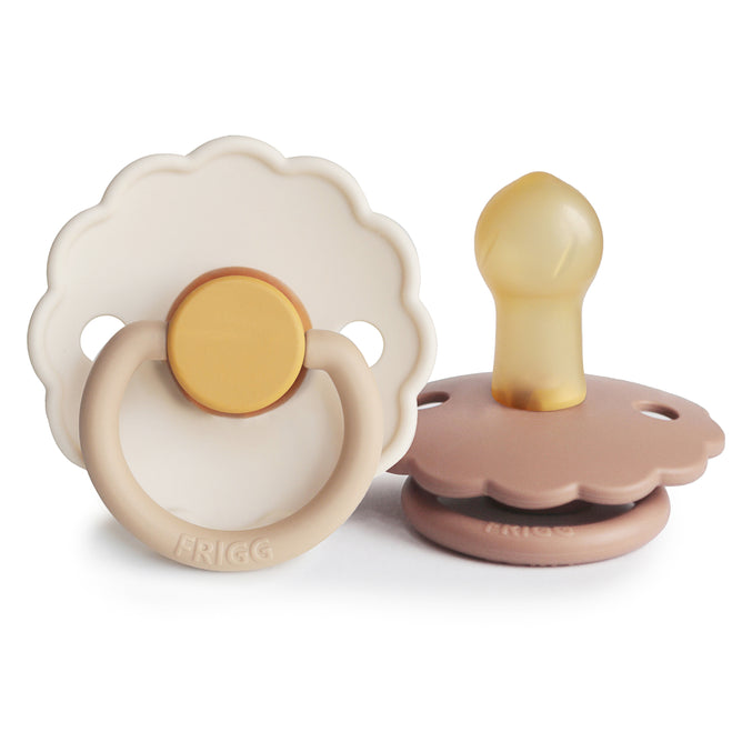 Two mushie FRIGG Daisy Natural Rubber Pacifiers (BPA-Free) are shown: one upright with a beige/yellow handle, the other lying down to reveal the silicone nipple and brown base. Both feature distinctive FRIGG branding.