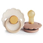Two mushie FRIGG Daisy Natural Rubber Pacifiers (BPA-Free) are shown: one upright with a beige/yellow handle, the other lying down to reveal the silicone nipple and brown base. Both feature distinctive FRIGG branding.