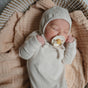 A newborn sleeps in a woven basket lined with a soft beige blanket, wearing a cream onesie and bonnet, holding a mushie FRIGG Daisy Natural Rubber Pacifier – BPA-Free in its mouth.
