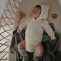 A baby wearing a knitted bonnet, cream onesie, and booties sleeps on a blanket in a wicker bassinet, with a wooden toy and the mushie FRIGG Daisy Natural Rubber Pacifier – BPA-Free nearby.