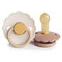 Two mushie FRIGG Daisy Natural Rubber Pacifiers (BPA-Free) are displayed: one stands upright showing its brown base and nipple, while the other lies flat revealing a beige and gold handle and a shield embossed with FRIGG.
