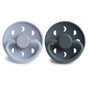 Two FRIGG Daisy Natural Rubber Pacifiers by mushie, BPA-free, in light and dark gray. Each has a circular shield with ventilation holes, a handle, and the embossed brand name for stylish, safe comfort.