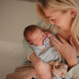 A woman lovingly cradles her sleeping newborn wrapped in a blanket. The baby is soothed with the mushie FRIGG Daisy Natural Rubber Pacifier – BPA-Free, while the woman smiles gently, holding her child close.