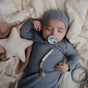 A baby in a gray outfit sleeps on a soft beige blanket with a mushie FRIGG Daisy Natural Rubber Pacifier – BPA-Free clipped to their clothes. Nearby are a plush star and teething ring, creating a peaceful scene.