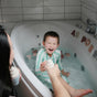 A young boy laughs in the tub filled with water and mushie Foam Letters & Numbers Bath Play Set – Educational, Water-Safe, while an adult shows him a toy ice cream cone.