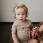 A light-skinned, curly red-haired baby in a beige floral onesie and headband sits on a dark wood floor, holding the mushie Silicone Pacifier Clip | Hera in pink. Another child’s hand is partially visible beside them.