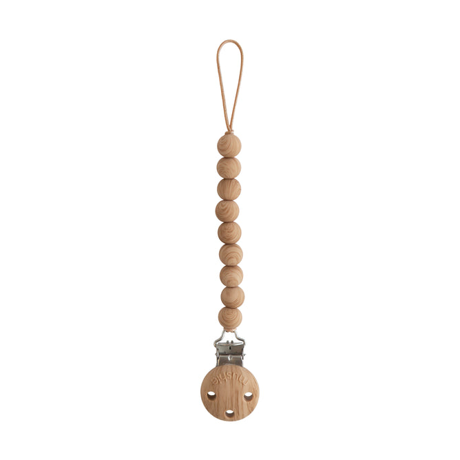 A baby essential, the mushie Silicone Pacifier Clip | Hera features round silicone beads on a cord, with a loop for attaching a pacifier and a metal clasp secured to a wooden disk at the other end.