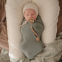 A newborn lies on neutral paisley bedding, wrapped in a sage green blanket, wearing a cream hat and using the mushie Silicone Pacifier Clip | Hera—one of the must-have baby essentials.