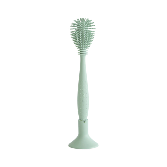 The mushie Silicone Baby Bottle Brush – Dual-Head, Non-Scratch, BPA-Free features a light green ergonomic handle with grip dots, soft silicone bristles, a textured round cleaning head, and comes with a matching upright stand.