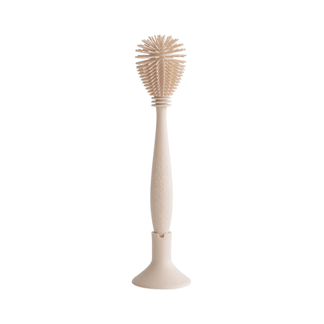 The mushie Silicone Baby Bottle Brush – Dual-Head, Non-Scratch, BPA-Free—shown in beige with an ergonomic handle—stands upright in a matching holder. Its rounded silicone bristles ensure effective cleaning against a white background.