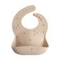 The mushie Silicone Baby Bib – Adjustable features a beige design with a small bee pattern, food-catching pocket, and adjustable snap neck closures. Made from FDA-tested food-grade silicone, it ensures safe and easy mealtimes for your baby.