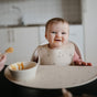 A smiling baby sits in a highchair wearing the mushie Silicone Baby Bib – Adjustable, made from FDA-tested food-grade silicone. A bowl and spoon rest on the tray, as an adult offers a spoonful. The kitchen sink is visible in the background.