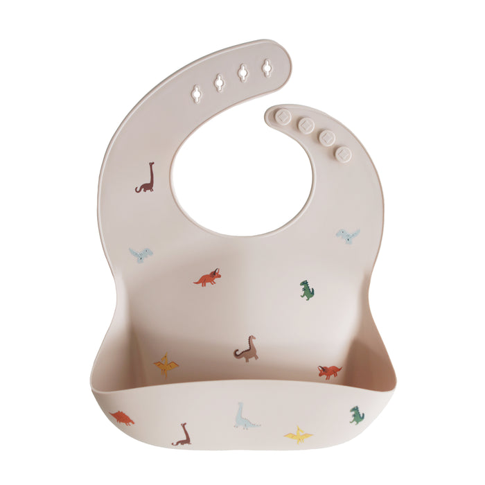 The mushie Silicone Baby Bib – Adjustable is a beige FDA-tested food-grade silicone bib with a pocket, colorful dinosaur prints, and an adjustable neck strap with four button fasteners.