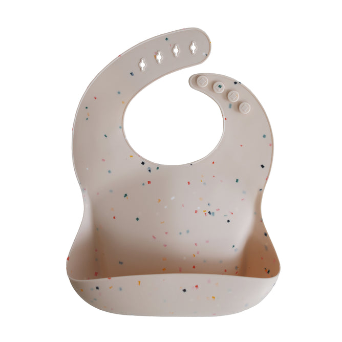 The mushie Silicone Baby Bib – Adjustable in light beige is made from food-grade silicone with a curved neckline, snap buttons, deep front pocket, and a playful scattered confetti dot pattern.