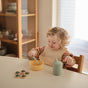 A young child with curly hair smiles at a table, eating from a yellow bowl. Nearby are mushie baby essentials, including the mushie Silicone Baby Bib – Adjustable, a green bottle, and a toy. Shelves with dishes appear in the background.