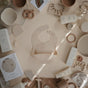 Flat lay of neutral mushie baby essentials on a beige background, highlighting the Silicone Baby Bib – Adjustable at the center, made from FDA-tested food-grade silicone, surrounded by bottles, plates, teethers, pacifiers, and more.