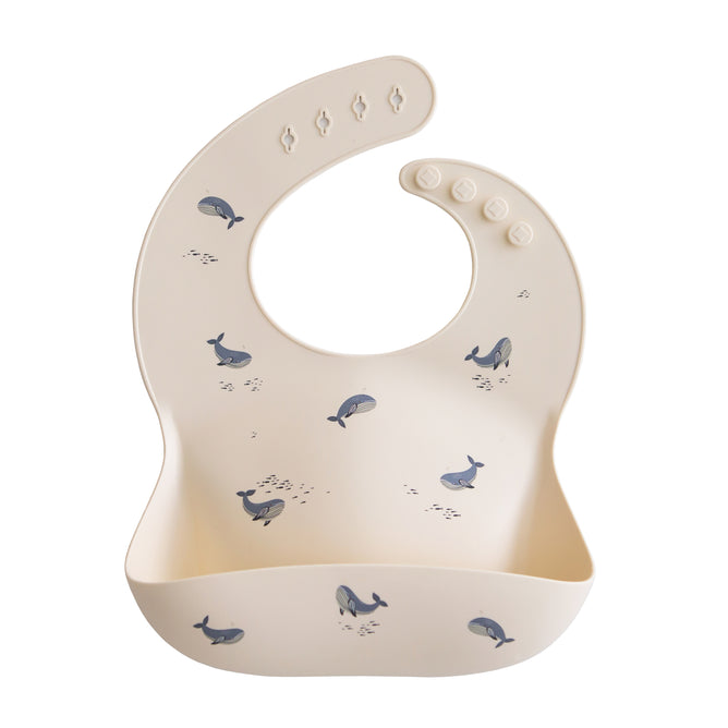 The mushie Silicone Baby Bib – Adjustable features a beige design with blue whale illustrations, a large food-catching pocket, and adjustable buttons. It’s made from FDA-tested, food-grade silicone for safe, mess-free meals.