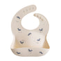 The mushie Silicone Baby Bib – Adjustable features a beige design with blue whale illustrations, a large food-catching pocket, and adjustable buttons. It’s made from FDA-tested, food-grade silicone for safe, mess-free meals.