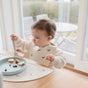 A toddler in a mushie Silicone Baby Bib – Adjustable sits by a window, eating from a blue divided plate. Patio furniture and greenery outside the window highlight the comfort of mushie baby essentials.