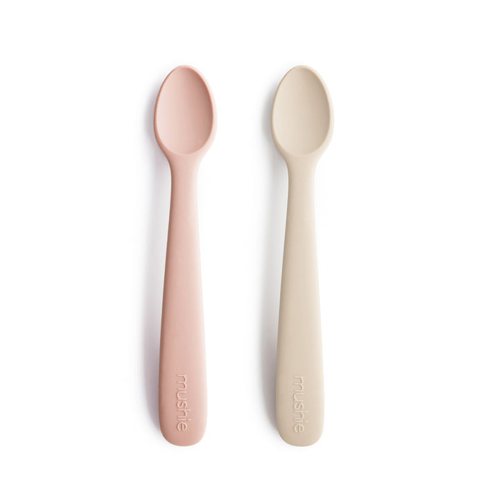 Two BPA-free, food-grade silicone feeding spoons by mushie—one pink, one off-white—lie side by side on a white background. Each features a rounded head and the brand name embossed on the handle.