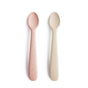 Two BPA-free, food-grade silicone feeding spoons by mushie—one pink, one off-white—lie side by side on a white background. Each features a rounded head and the brand name embossed on the handle.