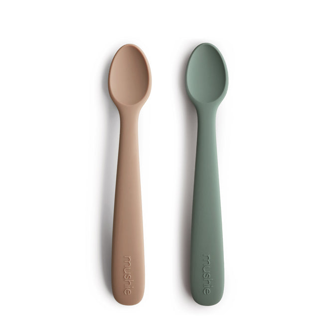 Two BPA-free, food-grade silicone feeding spoons from mushie in beige and sage green are shown side by side on a white background, each featuring the embossed mushie brand name on their handles.