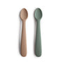 Two BPA-free, food-grade silicone feeding spoons from mushie in beige and sage green are shown side by side on a white background, each featuring the embossed mushie brand name on their handles.