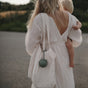A woman in a flowing white dress carries a small child and a cream-colored tote bag with a mushie Silicone Pacifier Holder Case attached, standing on a road with greenery in the background.
