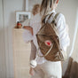 A woman with a tan Fjällräven backpack and a mushie Silicone Pacifier Holder Case carries a small child on her hip in a softly lit, minimalist room, wearing light-colored casual clothes—ideal for carrying baby essentials.