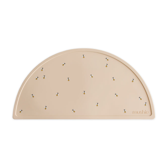 The mushie Silicone Placemat is a beige, semicircular mat made from 100% food-grade silicone, featuring small bee illustrations and the mushie logo embossed in the lower right corner—a delightful baby essential.