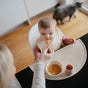 A baby wearing a bib sits in a high chair, being fed by an adult. On the tray is the mushie Silicone Placemat made from 100% food-grade silicone. Baby essentials are nearby, and a cat passes by a window in the background.