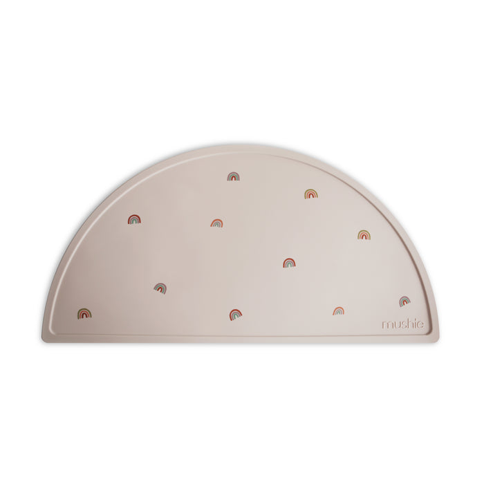 The mushie Silicone Placemat is a light beige, semicircular mat made from 100% food-grade silicone, featuring colorful rainbow designs and the embossed brand name—an ideal baby essential.