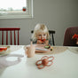 A young blonde child eats watermelon at a table set with mushie Silicone Placemat – 100% Food Grade Silicone, plates, a cup, and a teether. The child sits between red and pink chairs with a window in the background.