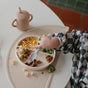A young child with a checkered sleeve uses a fork to eat macaroni, corn, broccoli, and meat from a divided plate on the mushie Silicone Placemat – 100% Food Grade Silicone, next to a beige sippy cup on a white table.