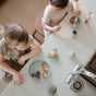 Two young children snack at a kitchen counter using mushie Silicone Placemats – 100% Food Grade Silicone – under their plates, with grapes and orange slices. Soft toys, baby items, and cups are also on the counter.
