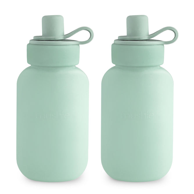 Two light green mushie Silicone Puree Pouches made from BPA-free, food-grade silicone with rounded shapes, matching caps, and loop handles are shown side by side on a white background.